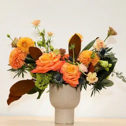 A speckled beige vase holds orange roses, yellow dahlias, and greenery, set against a plain light background—perfect flowers for sale in Manhattan NYC from your local flower shop.