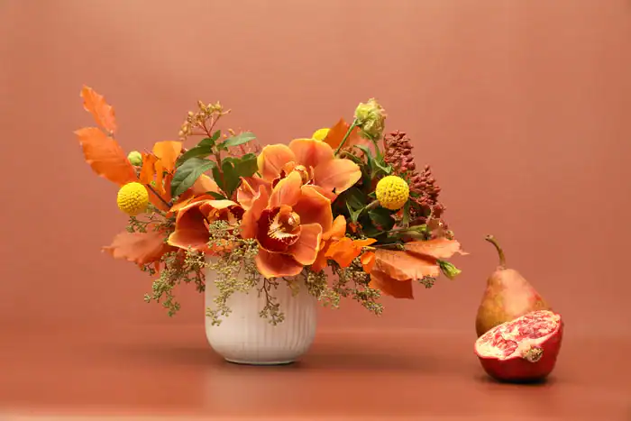 A white vase from a flower shop Manhattan NYC holds an autumn-themed arrangement with orange flowers, yellow spheres, and green leaves. Beside it sit a red pear and halved pomegranate, set against a rich brown background.