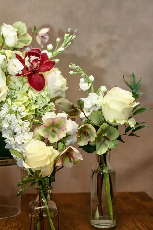 A floral arrangement featuring white roses, green and burgundy hellebores, and white hydrangeas in clear glass vases, set against a neutral background—perfect for your flower shop Manhattan NYC display.