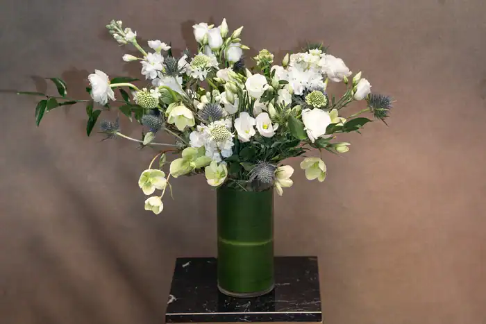 A green vase filled with an arrangement of white and light green flowers, including some thistle-like blooms, from a flower shop Manhattan NYC, displayed on a small black marble stand against a plain brown background.