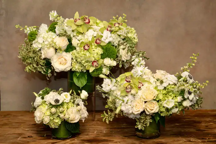 Three elegant floral arrangements featuring white and pale green flowers&mdash;including roses, hydrangeas, and orchids&mdash;are displayed in clear glass vases on a wooden surface. Beautiful options from a premier flower shop Manhattan NYC.