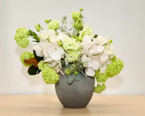 A gray ceramic vase holding an arrangement of light green and white flowers, including hydrangeas and lisianthus, sits on a light-colored surface&mdash;a perfect piece from a flower shop Manhattan NYC against a plain background.