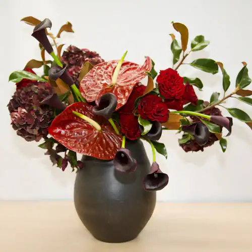 A black vase holds a modern floral arrangement with red anthuriums, dark red carnations, deep purple calla lilies, hydrangeas, and green foliage—perfect for those seeking striking flowers for sale in Manhattan NYC.