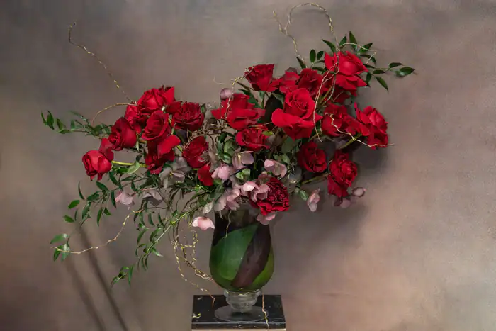 A glass vase filled with a lush arrangement of red roses, green leaves, and delicate pink flowers from a flower shop Manhattan NYC, set against a neutral, textured background.