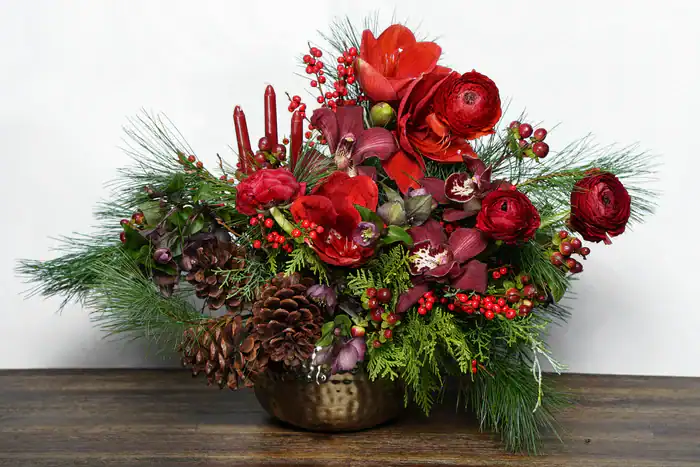 A festive floral arrangement in a brass bowl features red roses, ranunculus, pinecones, red berries, green pine branches, and red taper candles—perfect for anyone seeking flowers for sale Manhattan NYC. Displayed on a dark wooden surface against a white background.