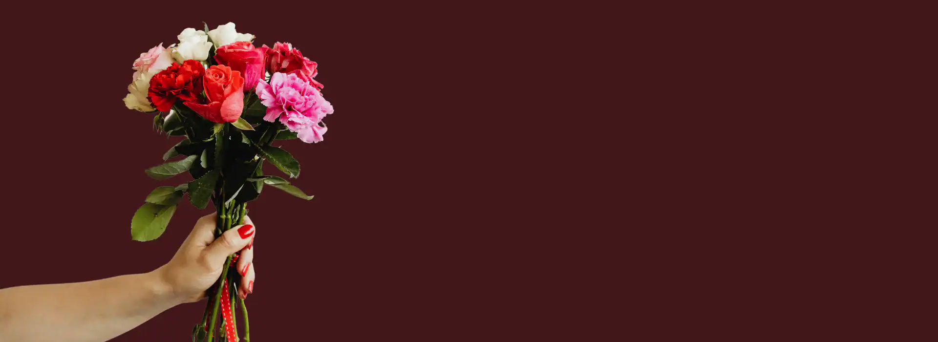 A hand with red-painted nails holds a bouquet of mixed flowers, including red, pink, and white roses from a flower shop Manhattan NYC, set against a plain dark maroon background.