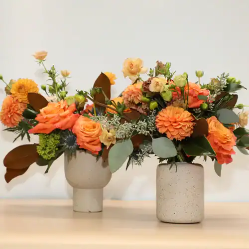 Two ceramic vases filled with vibrant orange, peach, and yellow blooms from a flower shop Manhattan NYC, accented by greenery and brown leaves, are displayed on a light wooden surface against a plain, light-colored background.