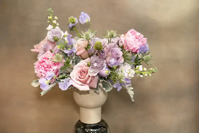 A ceramic vase holds a bouquet of pink and purple flowers, including roses, peonies, and greenery&mdash;an elegant arrangement reminiscent of blooms from a flower shop Manhattan NYC&mdash;set against a softly blurred, neutral background.