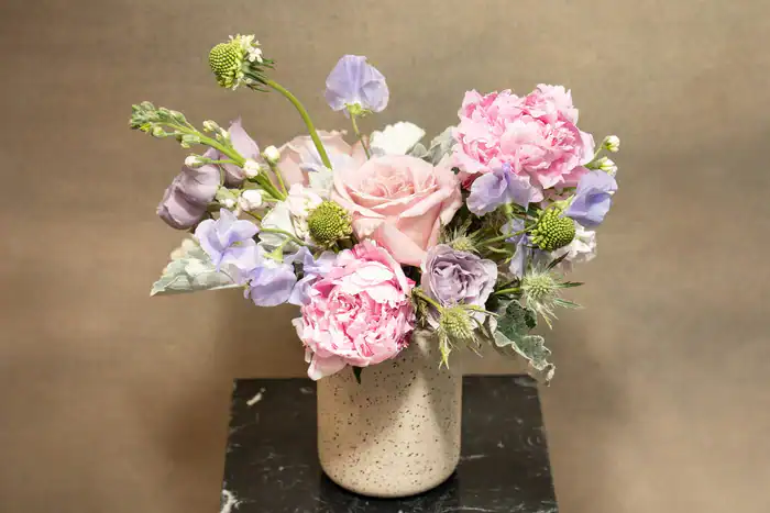 A speckled beige vase filled with pink peonies, pale purple roses, green buds, and silvery foliage sits on a black marble surface—a charming arrangement from a flower shop Manhattan NYC—against a plain taupe background.