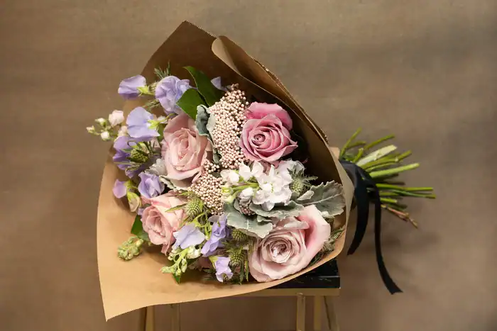 A bouquet of pale pink roses, lavender flowers, and greenery wrapped in brown paper with a black ribbon, resting on a wooden stool—perfect for those seeking flowers for sale in Manhattan NYC against a plain background.