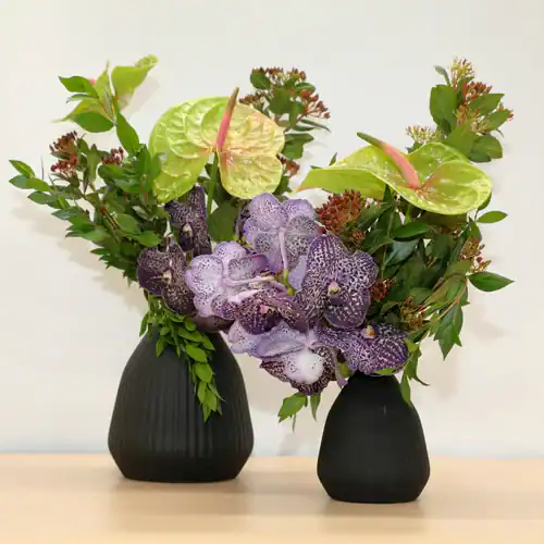 Two black vases hold arrangements of green leaves, pale green anthurium flowers with pink centers, and clusters of spotted purple orchids—an elegant display from our flower shop Manhattan NYC. Perfect for brightening any space.