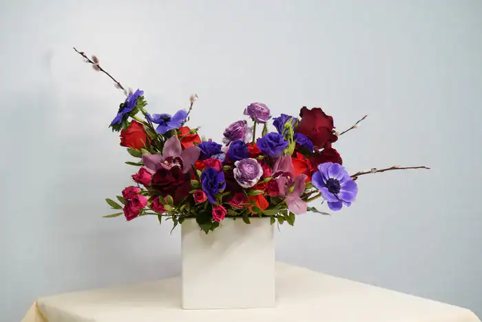 A white rectangular vase from a flower shop Manhattan NYC holds a colorful mix of red, purple, and violet flowers with green leaves and small branches, set on a light-colored surface against a pale blue background.