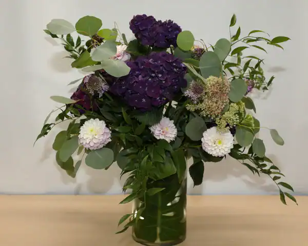 A glass vase filled with a mixed bouquet&mdash;large dark purple hydrangeas, pale pink dahlias, and green foliage&mdash;is displayed on a light wooden surface, perfect for those seeking flowers for sale in Manhattan NYC.