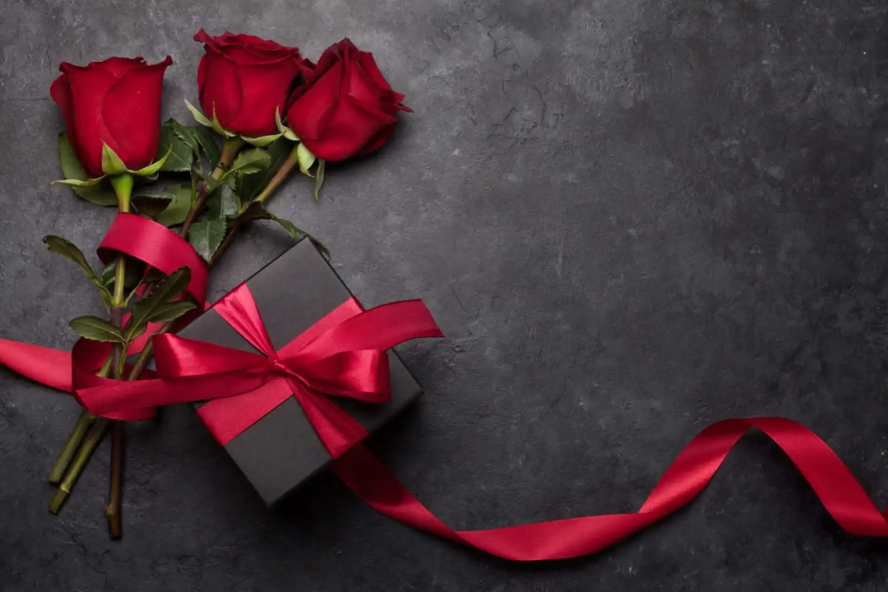 A black gift box with a red ribbon bow sits next to three red roses on a dark textured surface—an elegant display from a premier flower shop Manhattan NYC. The ribbon extends loosely to the right.