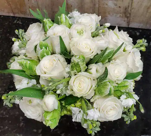 A bouquet of fresh white roses and greenery, arranged in a round shape with dewdrops on the petals, placed on a dark surface&mdash;a stunning option from a flower shop Manhattan NYC against a textured background.