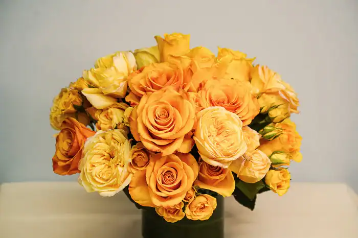 A bouquet of vibrant yellow and orange roses arranged in a green vase, set against a plain light blue background&mdash;perfect for anyone seeking flowers for sale Manhattan NYC.