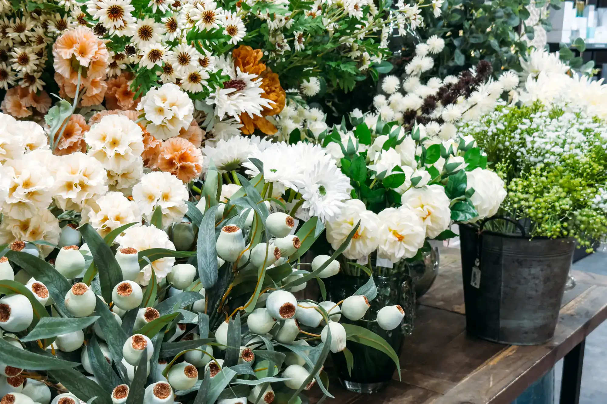 Buckets of assorted fresh flowers and greenery, including white peonies, daisies, and seeded eucalyptus, are displayed on a wooden table at a flower market.