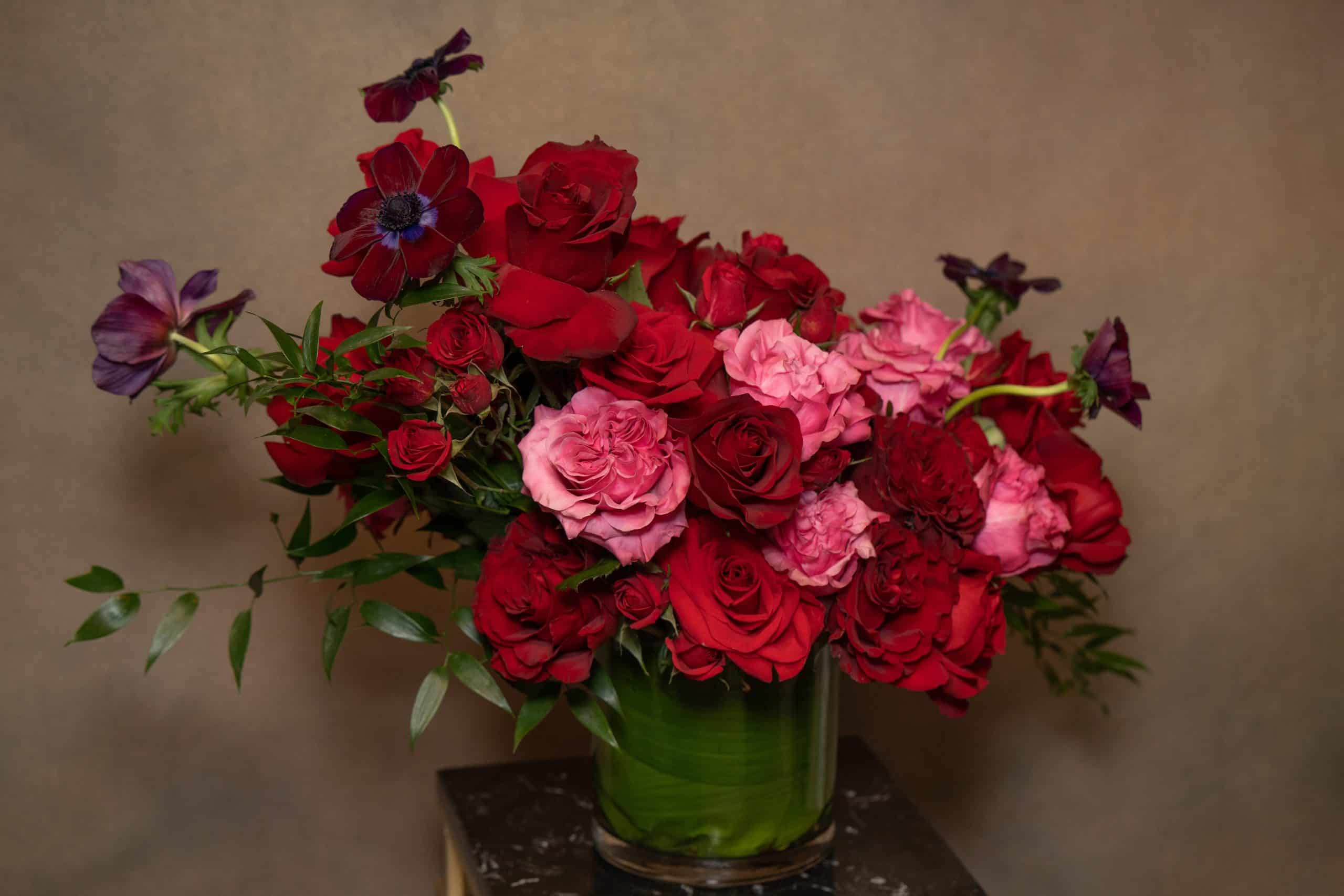 A glass vase filled with an abundant arrangement of red and pink roses, accented with dark purple flowers and green leafy stems, set against a neutral background.