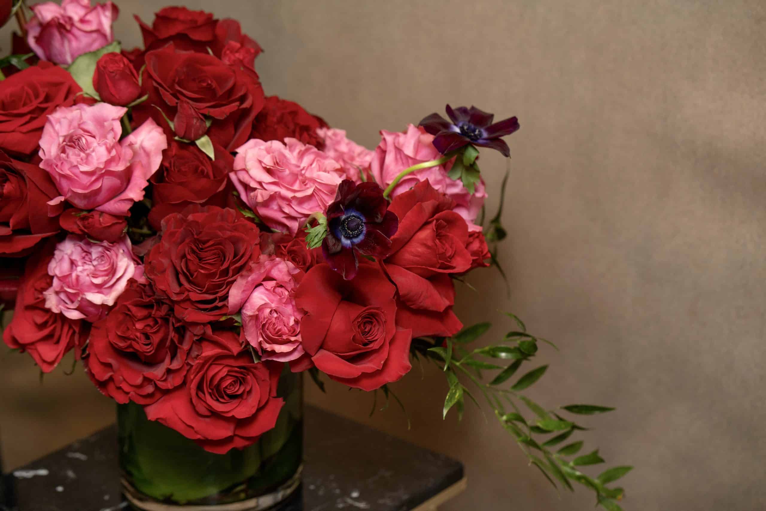 A bouquet of red and pink roses with a few dark purple flowers and green leaves arranged in a glass vase, placed on a black surface against a plain beige background.