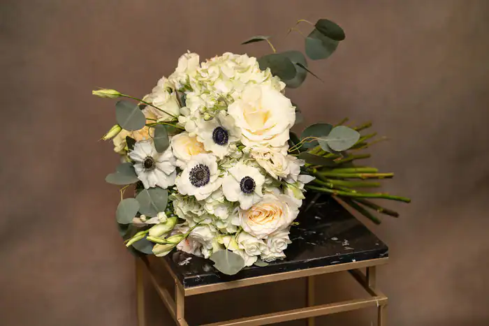A bouquet of white and cream flowers, including roses and anemones, with green eucalyptus leaves, rests on a marble-topped table against a brown background.
