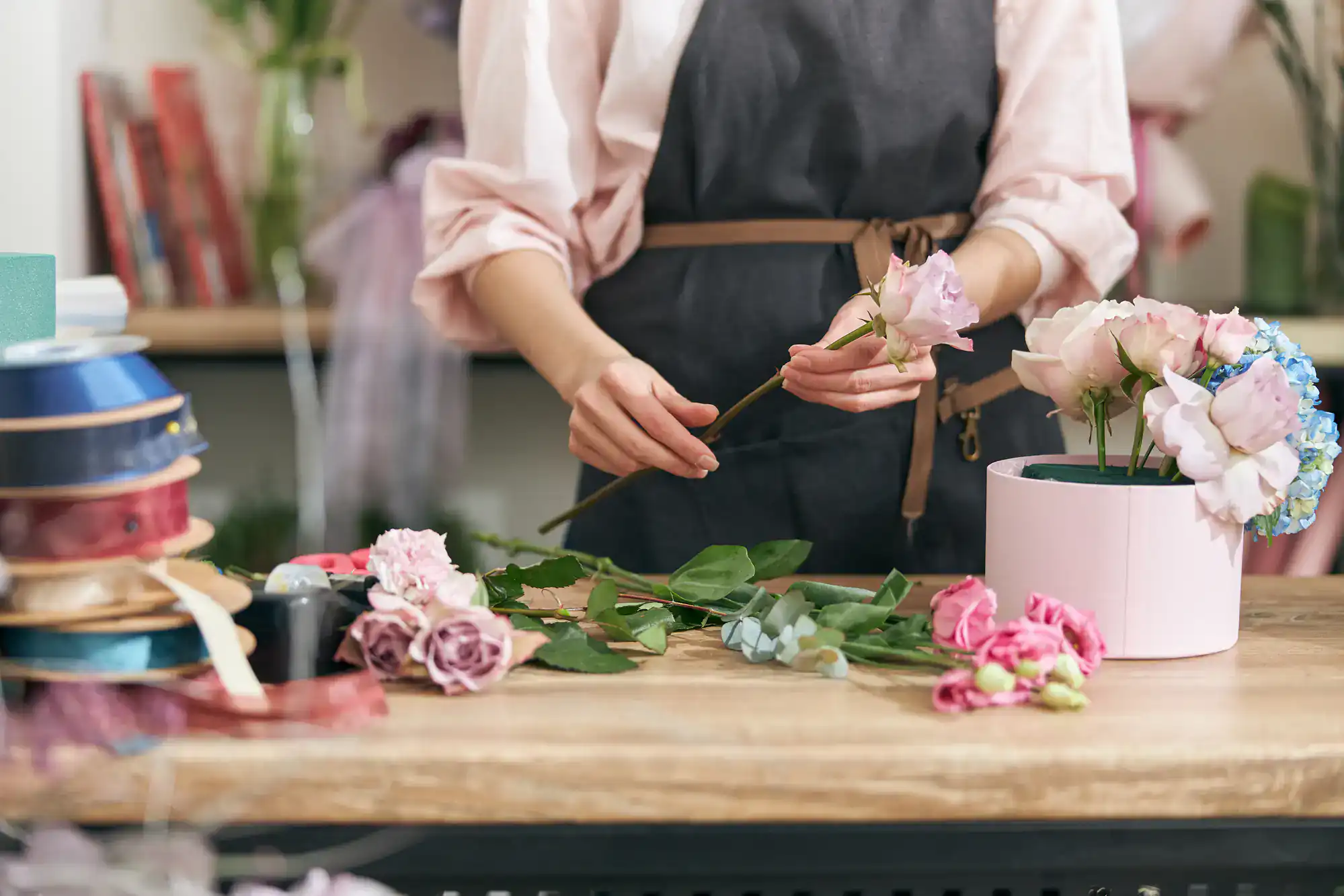 A person wearing a dark apron arranges pink and purple flowers on a wooden table, surrounded by ribbons and a round flower box. Only their torso and hands are visible.