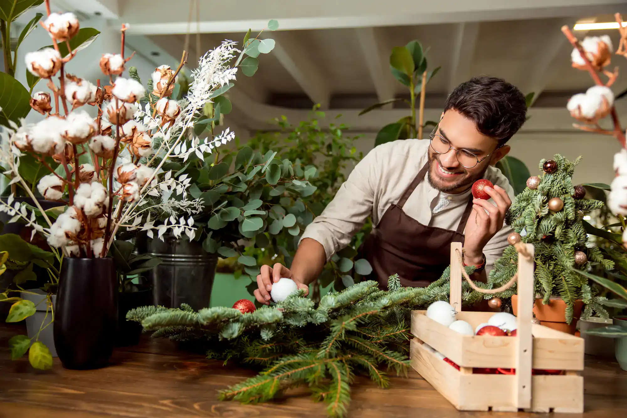 A smiling man wearing glasses and an apron decorates a pine branch with white and red ornaments at a wooden table, surrounded by various plants and festive decorations.
