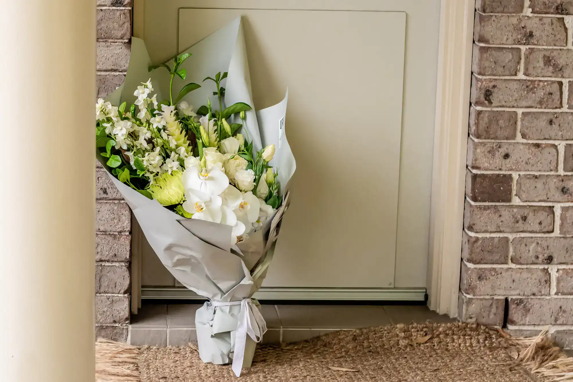 A bouquet of white and green flowers wrapped in gray paper is left on a doormat at a front door with a brick wall and beige pillar nearby.
