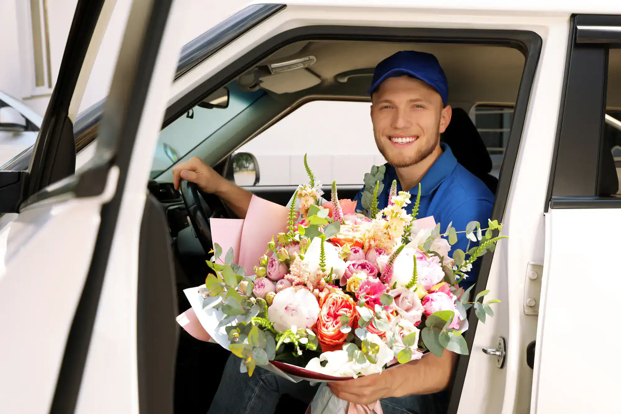A smiling delivery person in a blue cap and shirt sits in a car holding a large, colorful bouquet of flowers, ready to make a delivery.