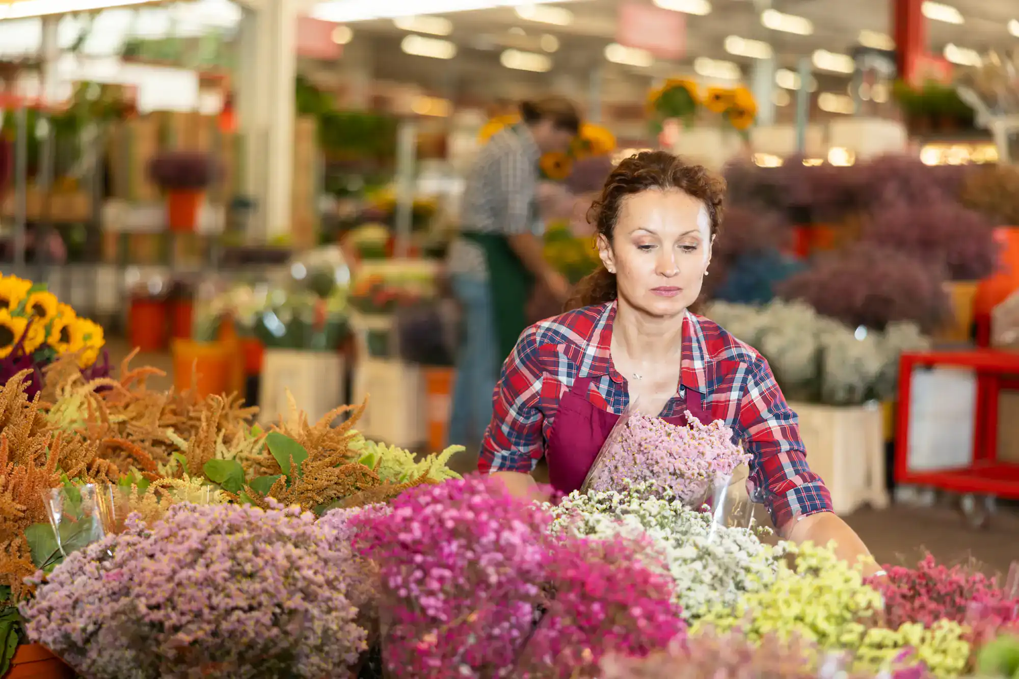 A woman in a plaid shirt and apron arranges an assortment of colorful flowers at a flower shop, with more flowers and another person visible in the background.