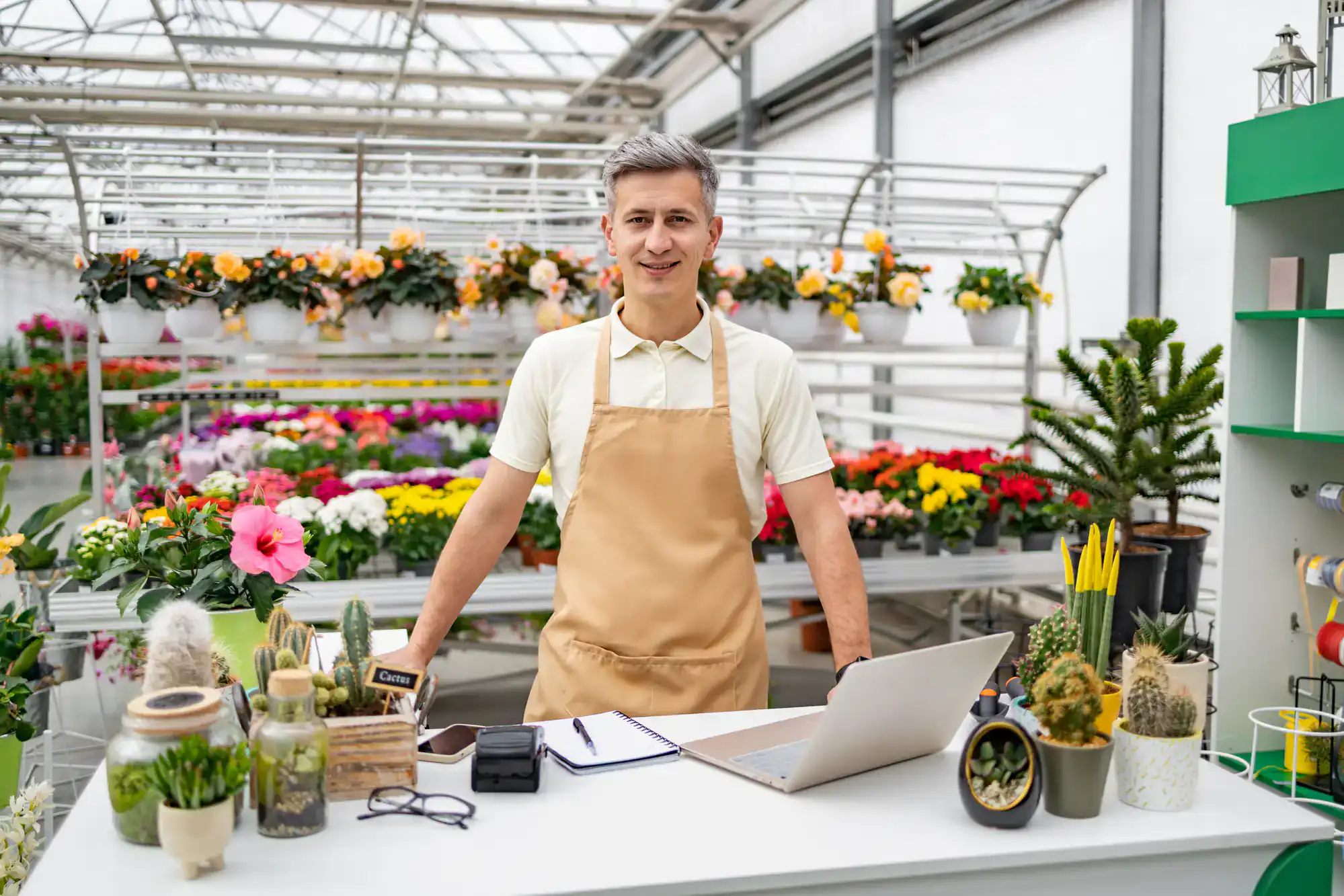 A smiling florist wearing a beige apron stands behind a counter in a greenhouse, surrounded by potted plants, cacti, flowers, a laptop, notepad, and gardening tools. Rows of colorful flowers are visible in the background.