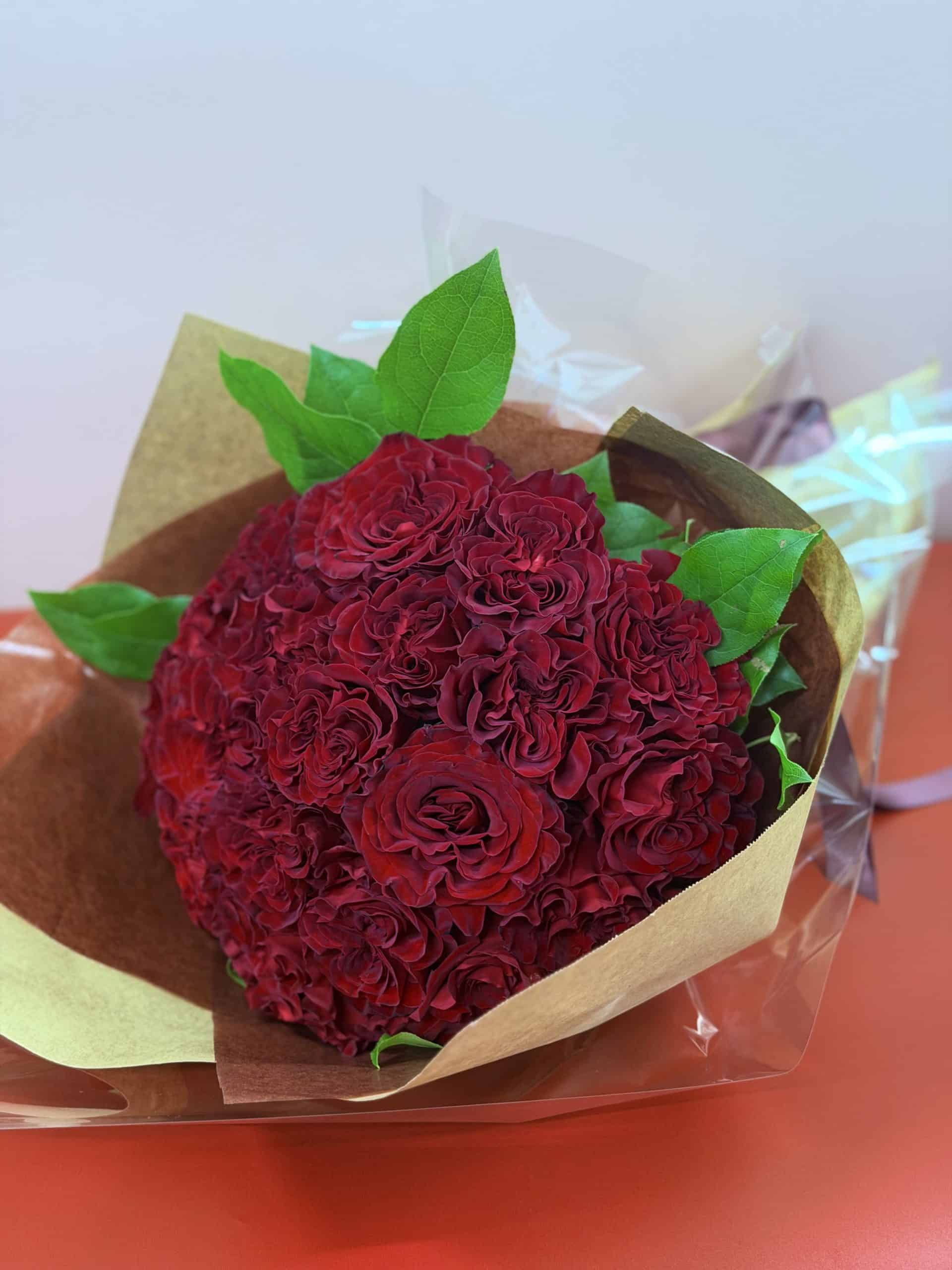 A bouquet of deep red roses and green leaves wrapped in brown paper and cellophane, placed on a red surface against a pale background.