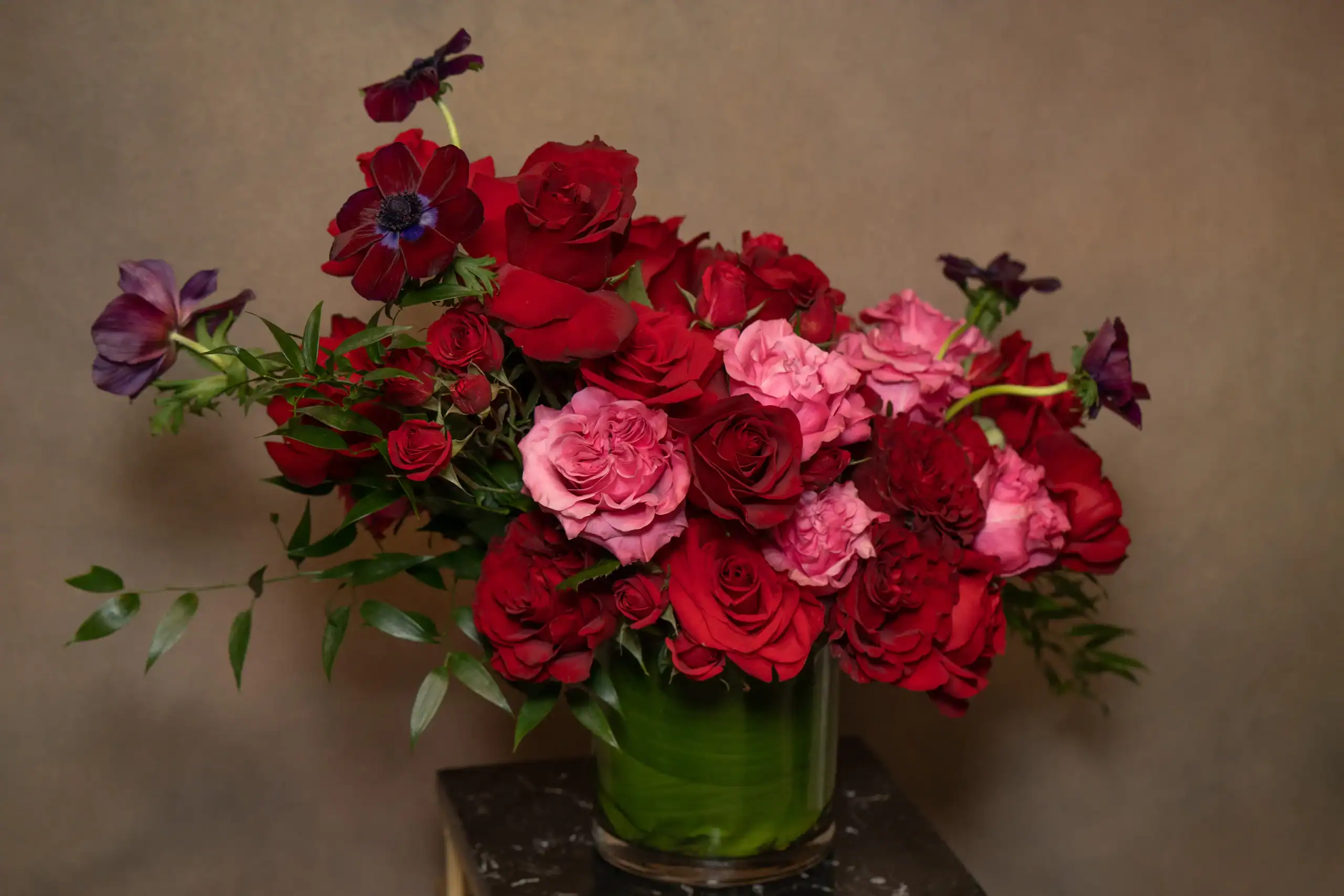 A lush bouquet of red and pink roses with a few deep purple flowers and green foliage arranged in a glass vase against a plain beige background.