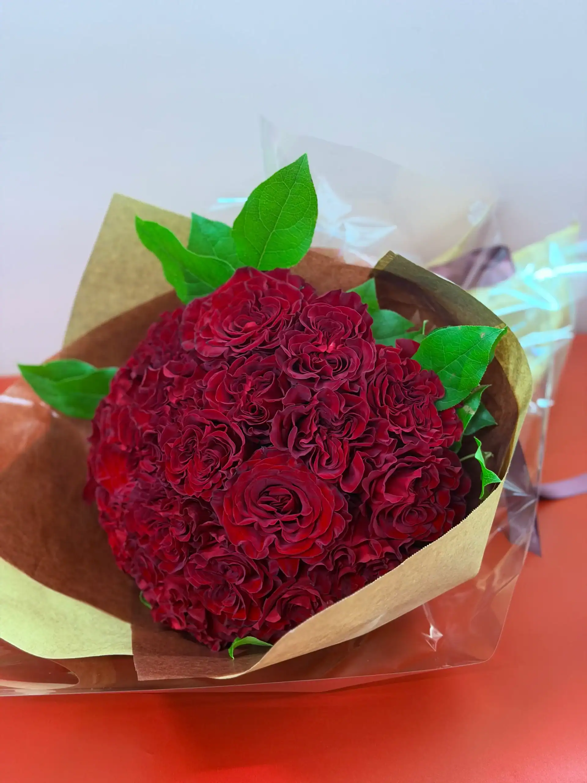 A bouquet of deep red roses with green leaves, wrapped in brown and transparent paper, sits on an orange surface.