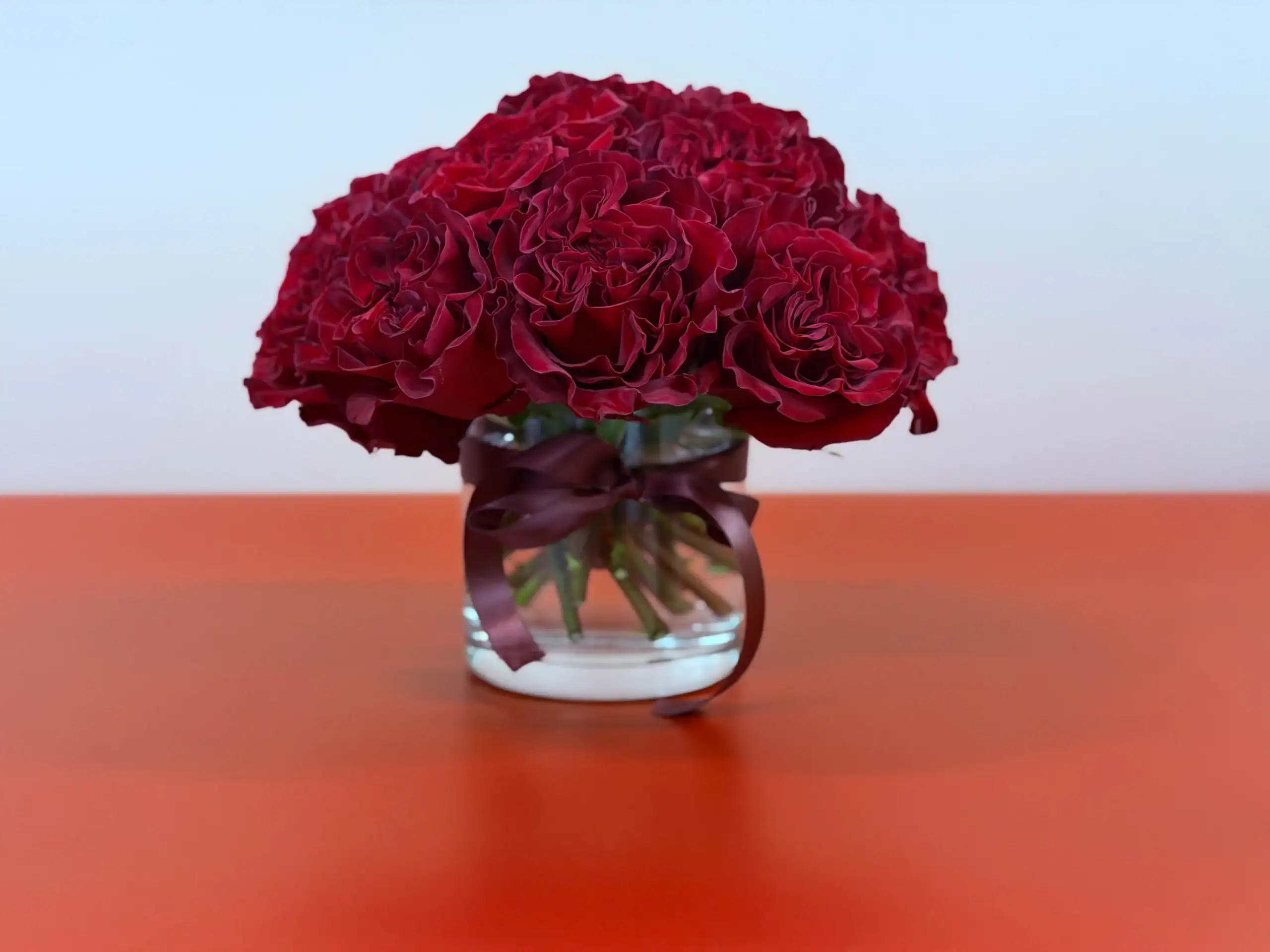 A clear glass vase filled with deep red roses, tied with a matching dark red ribbon, sits on an orange surface against a pale background.