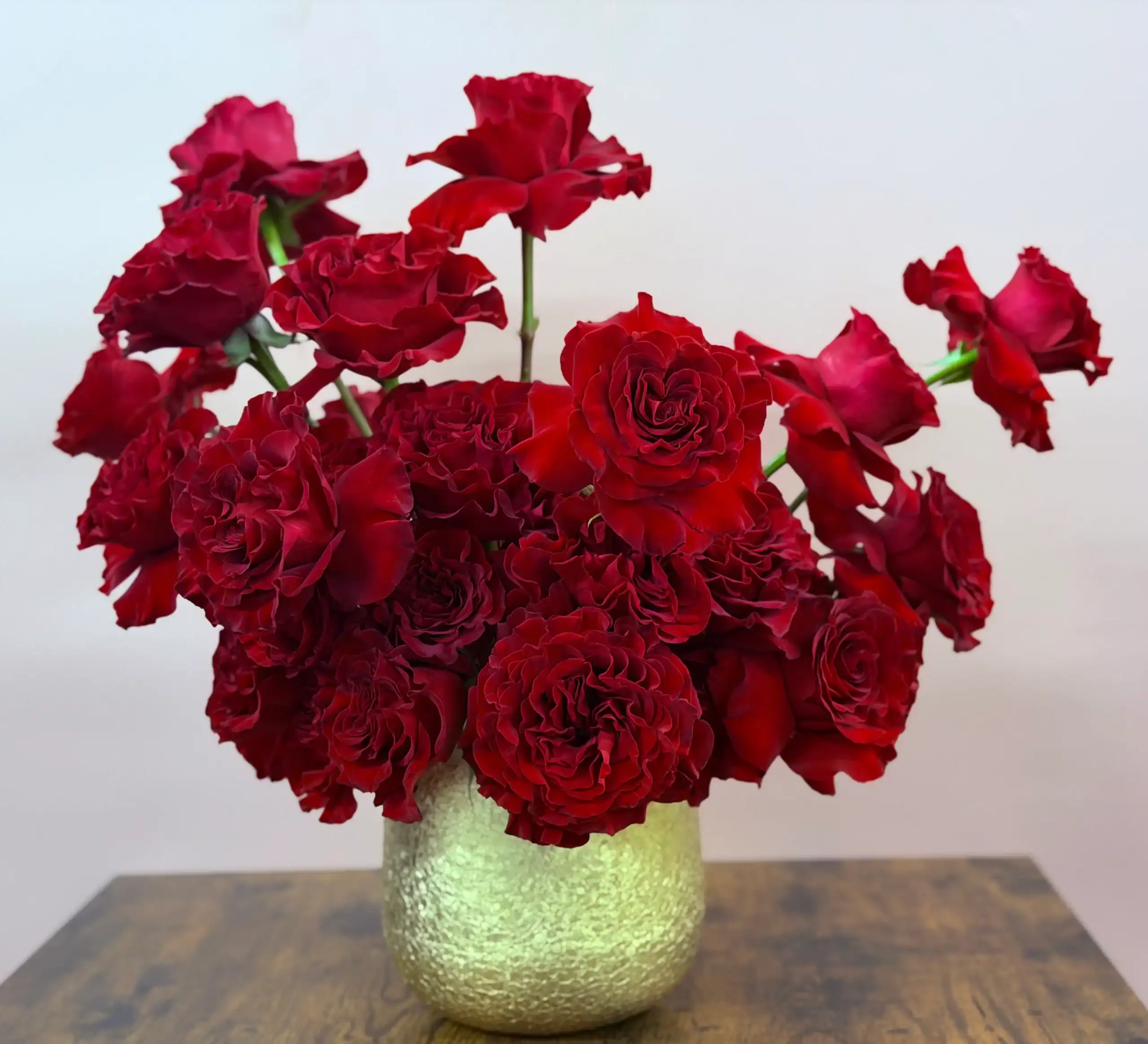 A gold textured vase filled with a bouquet of blooming deep red roses, placed on a wooden surface against a plain light-colored background.