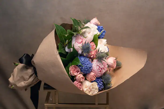 A bouquet of pink, white, and blue flowers with green leaves is wrapped in brown paper and tied with a black ribbon, resting on a gold-colored stool against a plain, neutral background.