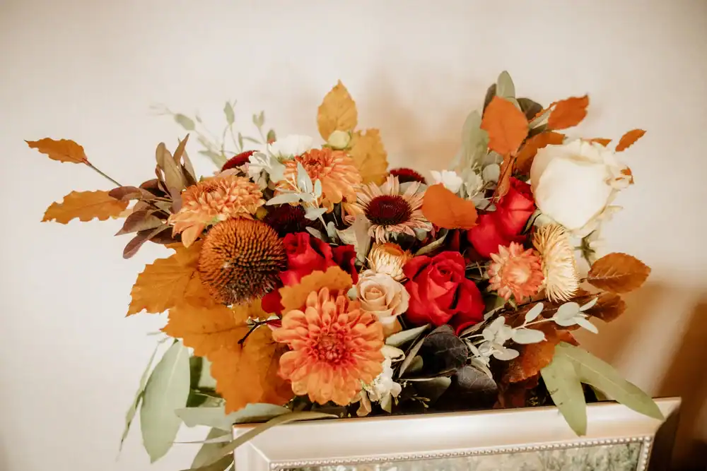 A bouquet of autumn-themed flowers featuring orange chrysanthemums, red and white roses, golden leaves, and greenery, arranged in a vase against a light-colored wall.