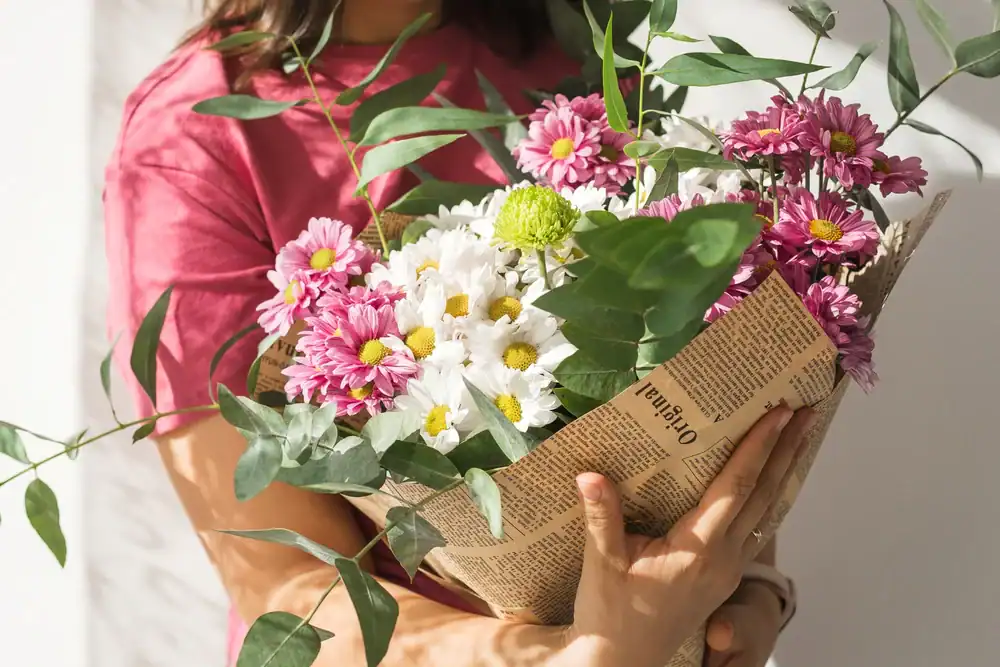A person in a pink shirt holds a bouquet of pink and white daisies with green leaves, wrapped in newspaper. Sunlight shines on the flowers and the person's arms.
