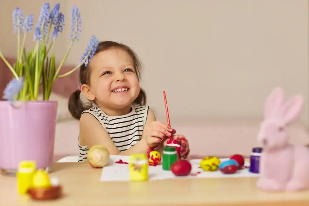 A smiling young girl in a striped shirt sits at a table, painting colorful Easter eggs. There are paint jars, decorated eggs, a pink rabbit figurine, and a potted flower on the table.