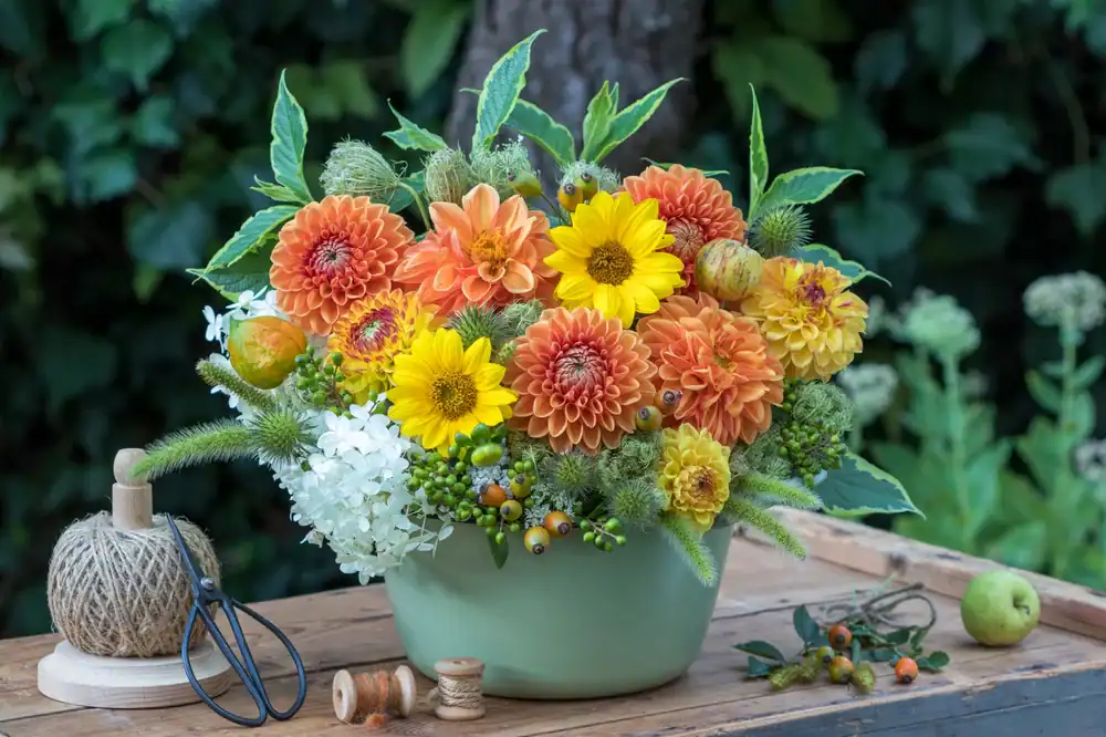 A green vase filled with yellow and orange flowers, green leaves, and small berries sits on a wooden table. Beside it are scissors, twine, and spools. A blurred leafy background completes the scene.