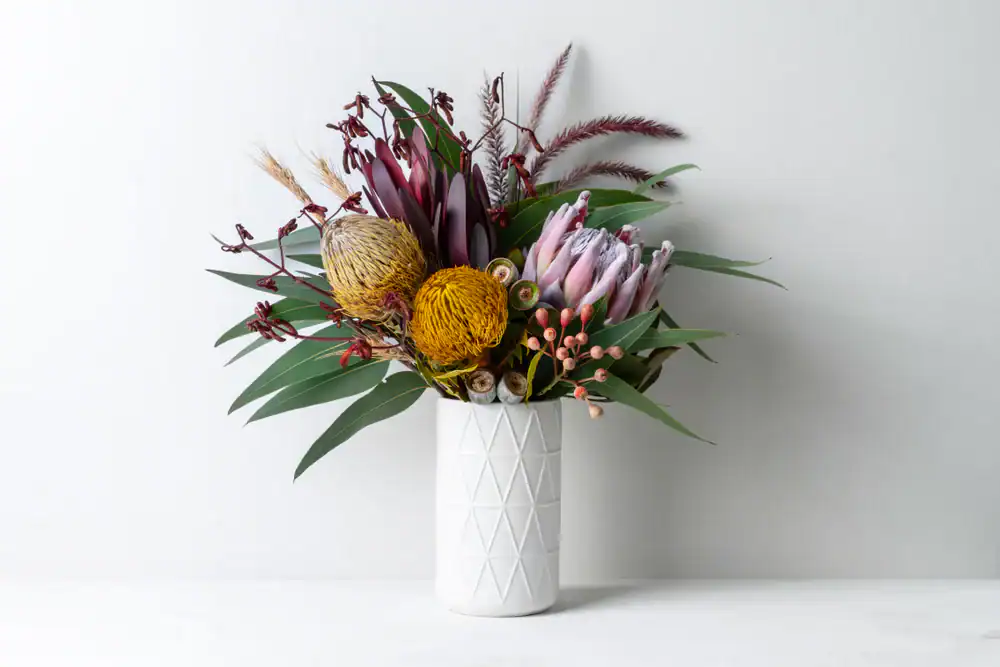 A modern white vase holds a vibrant arrangement of native Australian flowers, including protea, yellow billy balls, eucalyptus leaves, and pink berries, set against a plain white background.