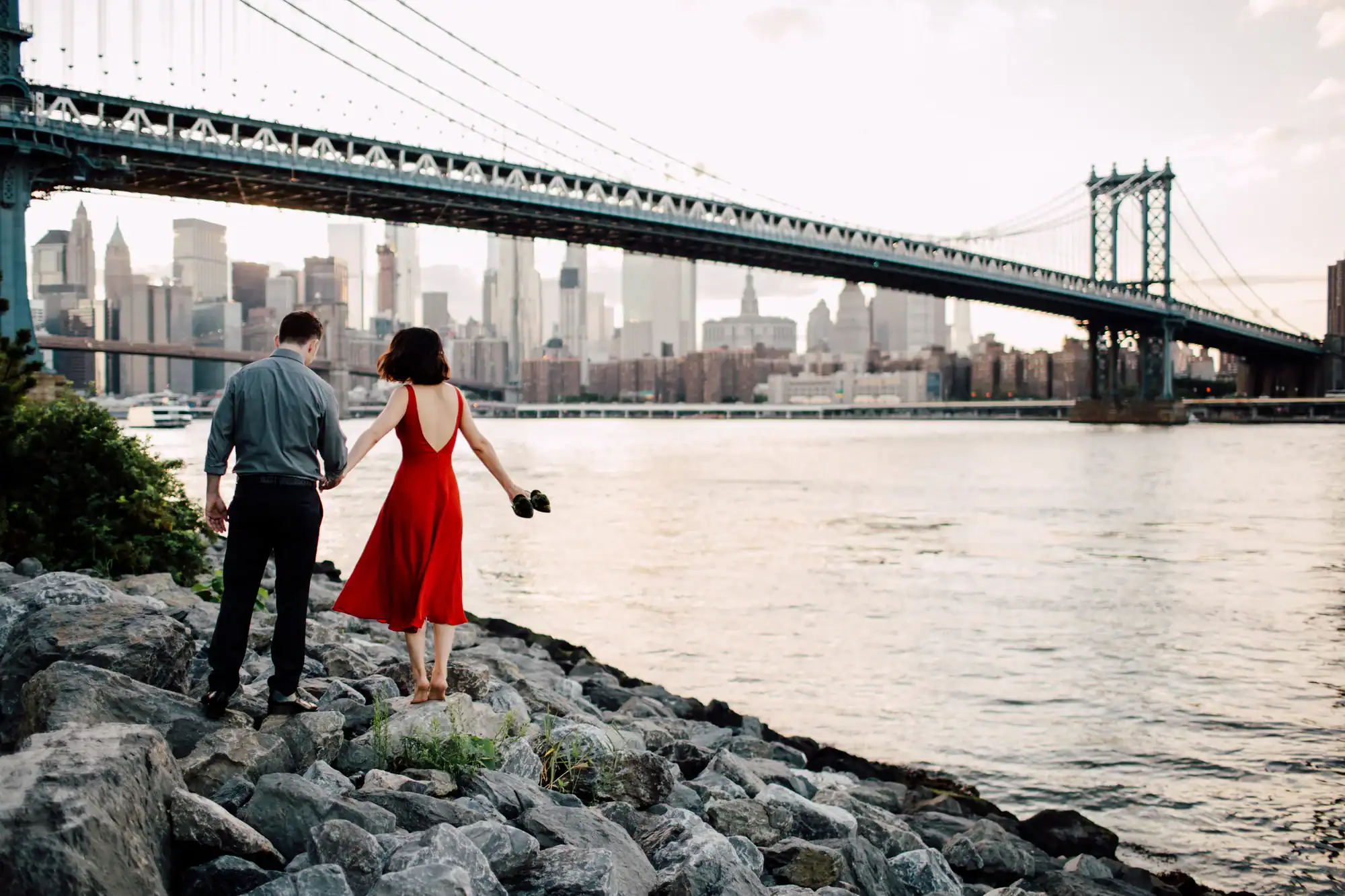 A couple walks hand in hand on rocky ground by a river, with the woman wearing a red dress. A large suspension bridge and city skyline are visible in the background.