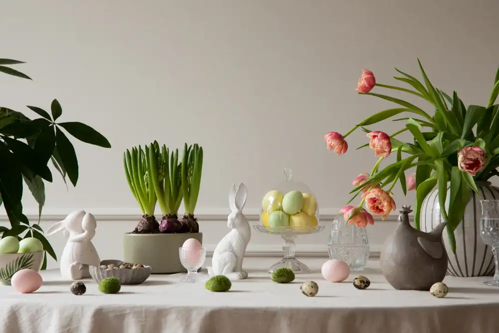 A decorated table features pastel eggs, white ceramic rabbits, blooming tulips in a vase, potted green plants, and glassware, creating a festive and spring-inspired centerpiece.