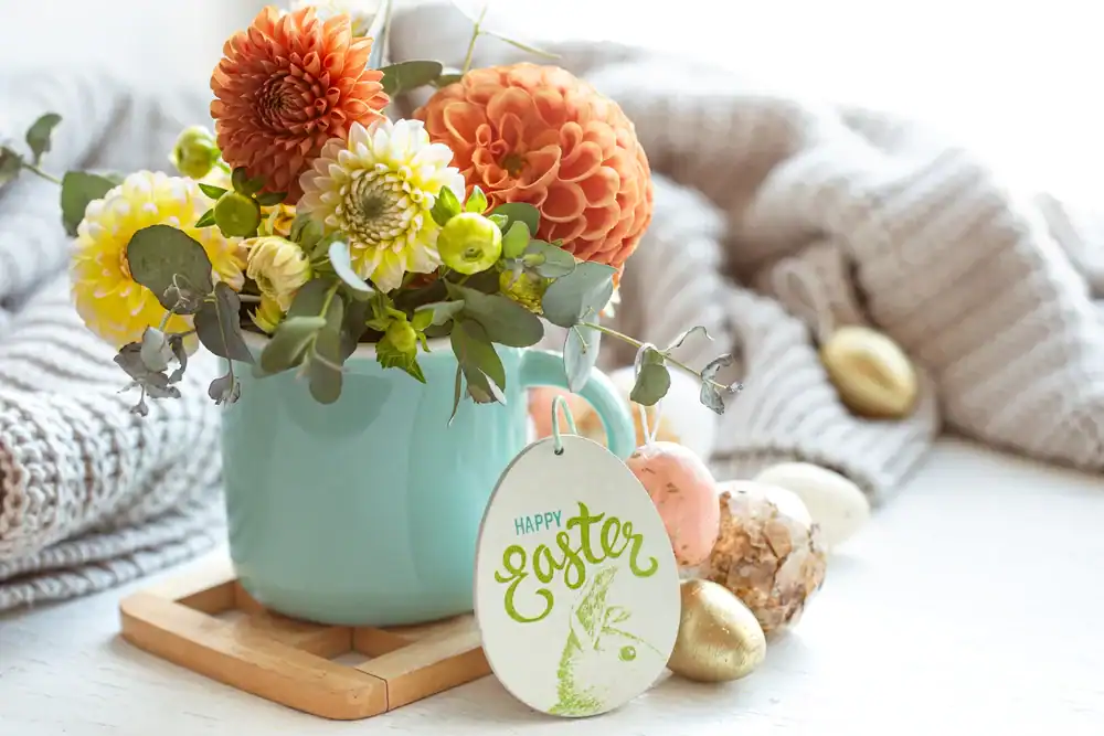 A blue vase holds orange and yellow flowers next to a wooden Easter decoration with "Happy Easter" and a bunny. Decorative eggs and a knit blanket are in the background.