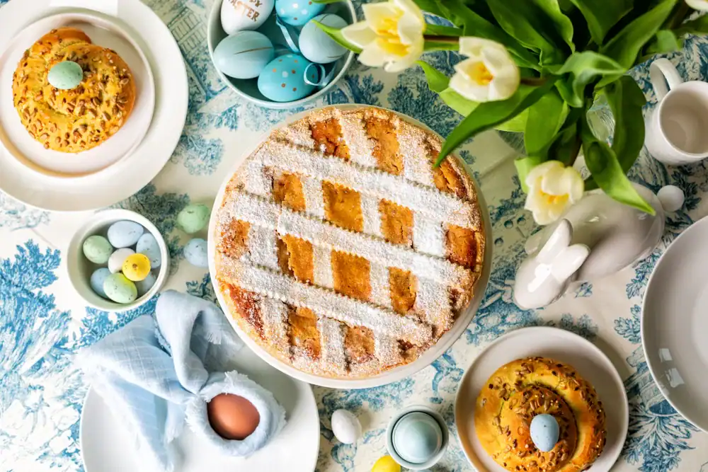 A table set for Easter with a lattice-topped pie, small cakes, pastel-colored eggs, a napkin-wrapped brown egg, white tulips in a vase, and decorative ceramic bunny on a blue and white tablecloth.
