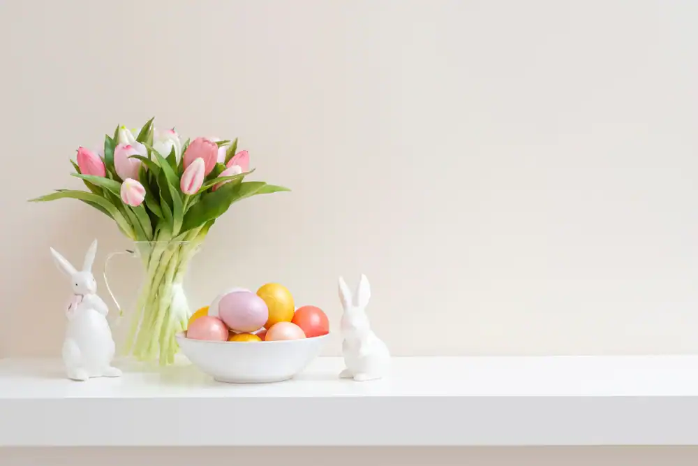 A white shelf displays a vase of pink tulips, a bowl of colorful Easter eggs, and two white ceramic bunny figurines against a plain light background.
