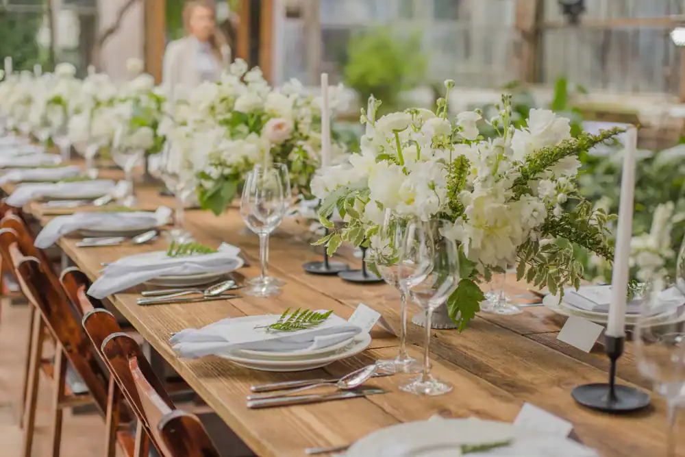 A long wooden table set for a formal event with white plates, napkins, wine glasses, silverware, white floral centerpieces, and white candles in holders; greenery surrounds the elegant setting.