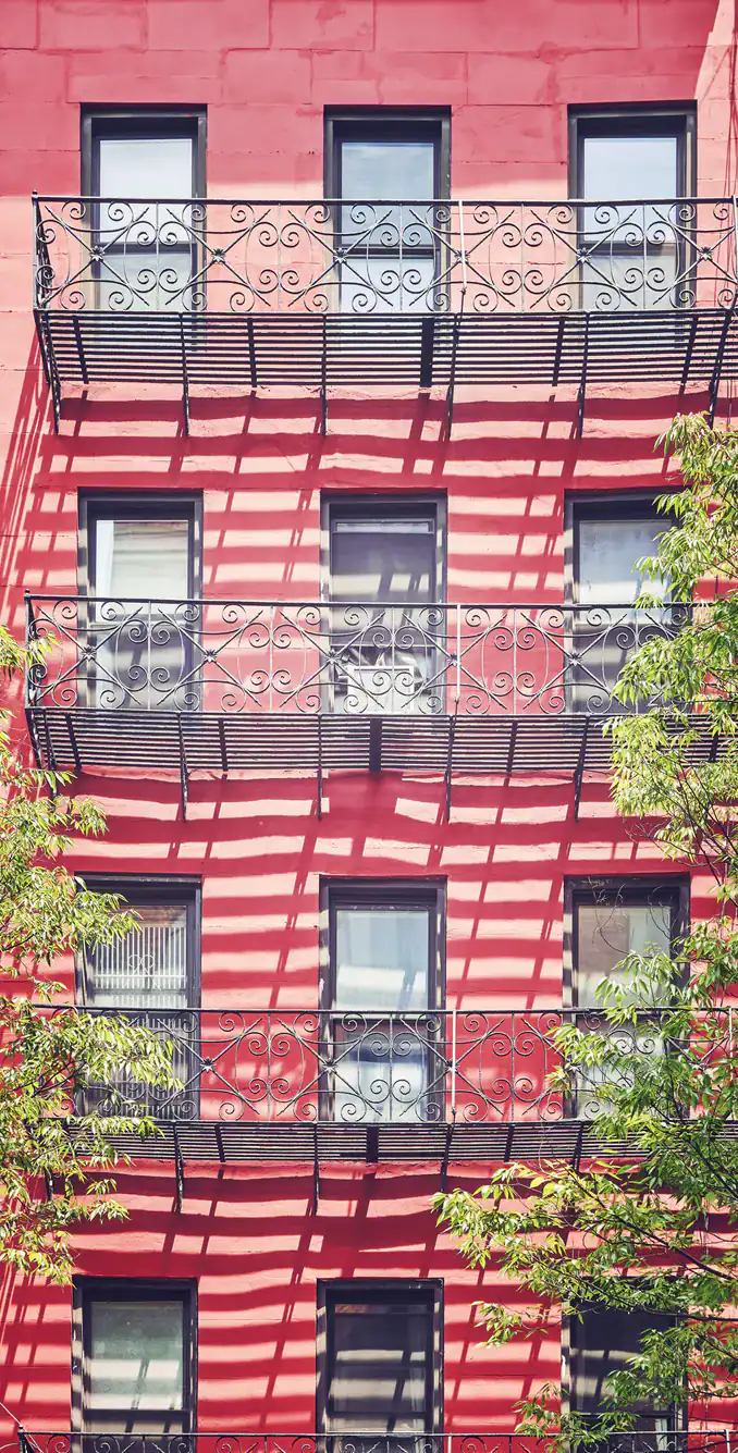 A red building with black wrought-iron balconies, casting striped shadows across the facade. Several windows are visible, and green tree branches frame the lower right and left corners.