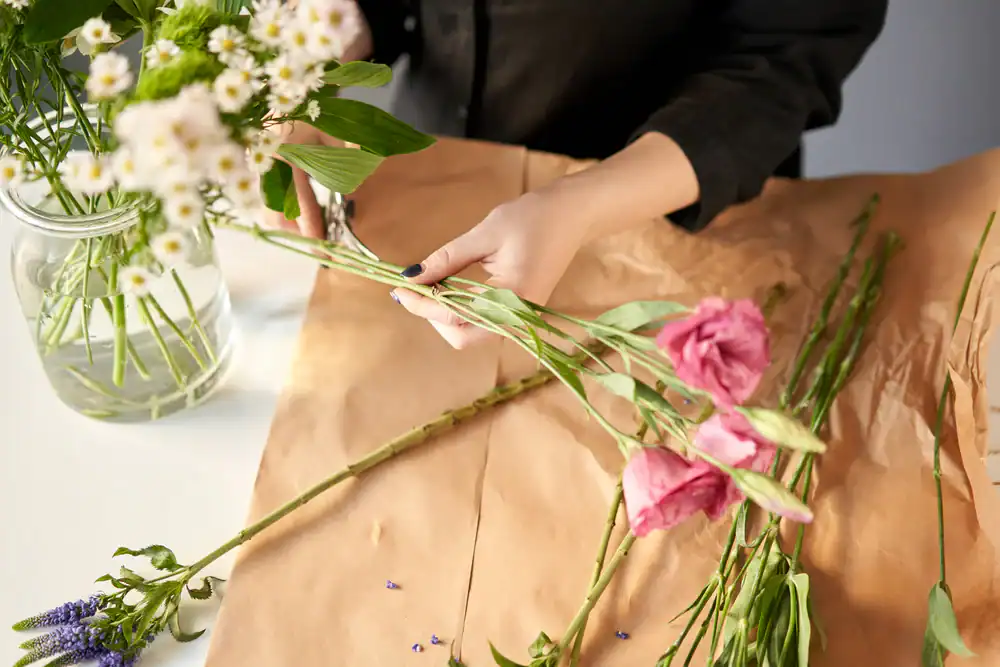 Person arranging a bouquet, holding and trimming pink flowers over brown paper. A clear jar with white flowers and greenery is nearby on the white table.
