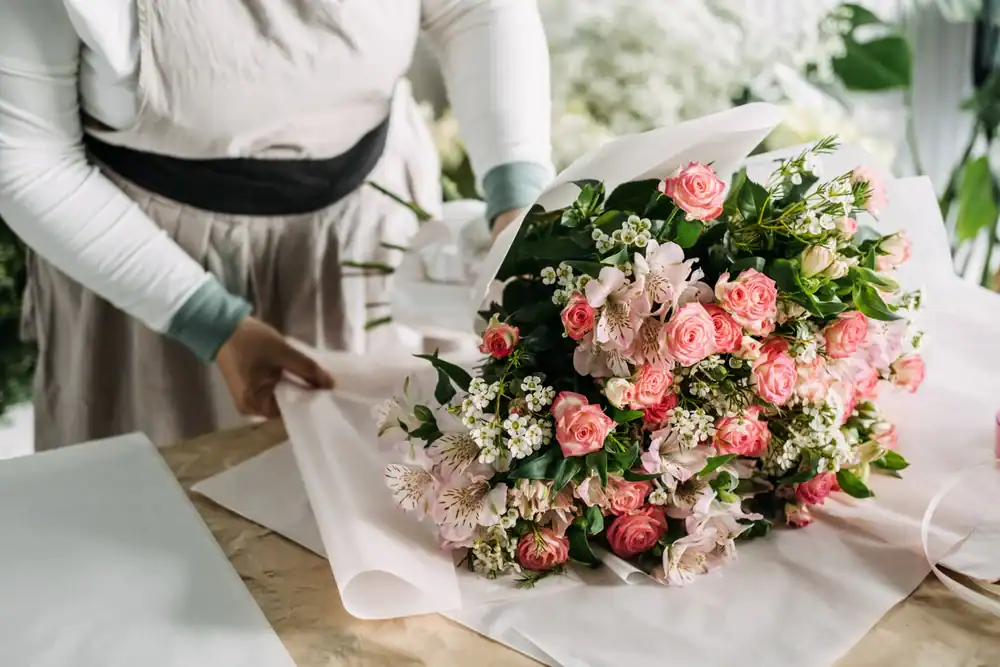 A person wrapping a bouquet of pink roses and white flowers in light pink paper on a table, wearing a light-colored long-sleeve shirt and an apron.